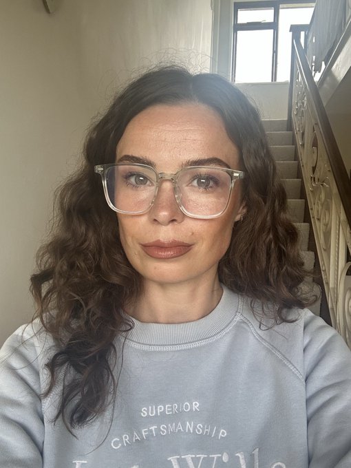 A woman with wavy brown hair and clear-framed glasses takes a selfie, wearing a gray sweatshirt with the text 'SUPERIOR CRAFTSMANSHIP' visible on the front, with a staircase and window in the background.