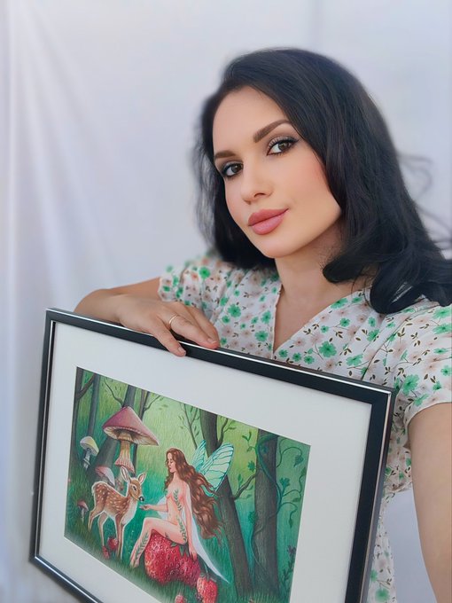 A woman with long dark hair and full makeup wearing a white floral dress, holding up a framed watercolor painting of a fairy sitting in the woods with a deer and a mushroom.