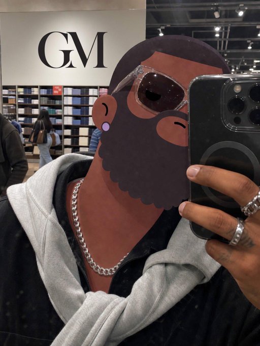 The image features a man taking a selfie with his cell phone, wearing sunglasses and a black shirt. He has a beard and is holding up his phone for the camera. The background of the photo shows a store or mall setting, indicating that they are in an indoor shopping environment. There are other people present in the scene as well, suggesting that it might be a busy day at the location where the man took the selfie.