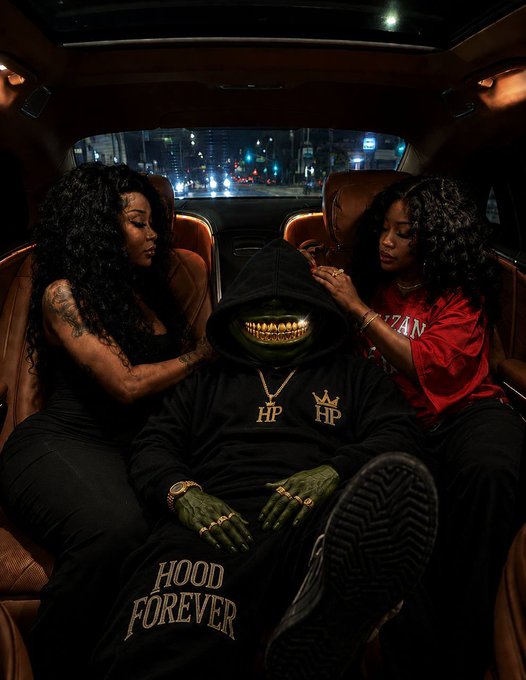 Two women sit in the back of a car flanking a hooded figure wearing gold teeth and 'HOOD FOREVER' pants with HP crown logos, with city lights visible through the rear window.