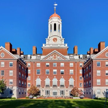 The image shows a stately brick building with a white clock tower topped by a weathervane, characteristic of the historic campus of Harvard University. The structure features symmetrical wings and is set against a clear blue sky with manicured lawns in the foreground, visually representing the institution mentioned in the headline about Harvard's $101 million Bitcoin ETF disclosure.