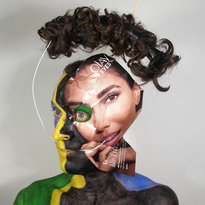 An avant-garde self-portrait or artistic photo featuring a person with body paint in Brazilian flag colors (yellow, green, blue) wearing a magazine mask over their face with the text 'OLAY EYES' and 'Make Me Like Me', topped with curly hair pieces.