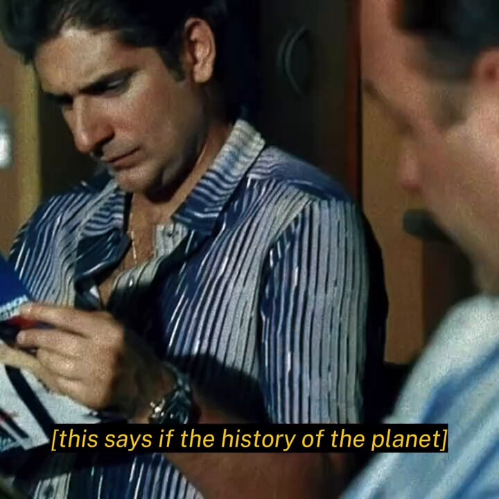 The image shows a man wearing a striped shirt, sitting down with his hands on his lap while reading a book or magazine. There are two other people partially visible at the bottom of the image, but they do not appear to be interacting with the main subject. The text in the image reads "This says if the history of the planet".