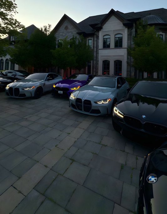 Several BMW cars, including a distinctive light blue M3 and a purple model, are parked on a brick driveway in front of a large stone mansion at dusk.