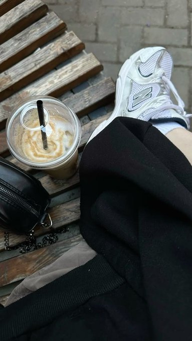 A first-person view of legs resting near a wooden bench, with an iced coffee drink, a small black purse, and a white sneaker visible.