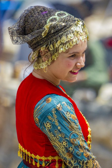 A smiling young woman wears a richly embroidered turquoise and crimson traditional garment, complemented by an ornate metallic headdress adorned with dangling gold coins and intricate filigree. The image visually embodies the "Colors of Heritage" theme, celebrating the vivid textiles and cultural craftsmanship highlighted in the Tezos NFT listing.