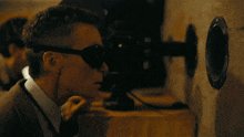 The image shows a man wearing sunglasses, sitting at a desk with his head tilted down towards a camera. He appears to be looking into the camera lens or focusing on something behind him. The background of the image features a wall that has a hole in it, and there is also another person partially visible in the top left corner of the photo. The caption reads "$FLR bombshell in 1 hour." This suggests that the man might have been involved in an event where he was required to take photos or videos using a camera with a built-in microphone.