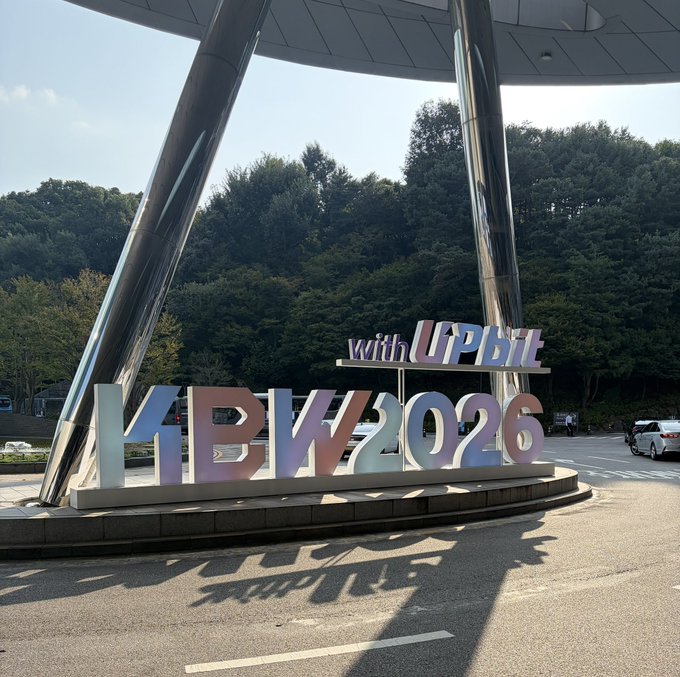 A colorful "with UPBEAT KFBW2026" sign is displayed beneath a modern architectural structure, set against a backdrop of lush green trees and a clear sky. The image relates to the post by visually representing the KBW 2026 event with UPBEAT, capturing the stabilization and branding moment for @kbwofficial.