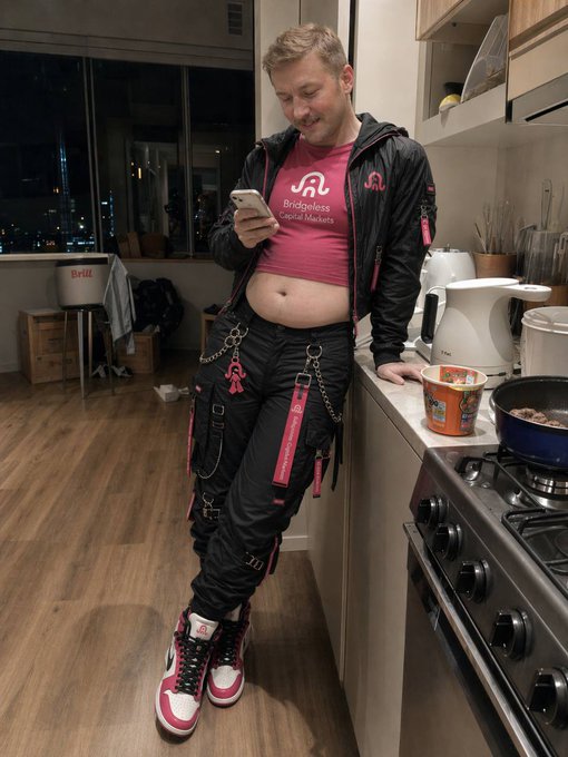 A man wearing a pink shirt with "Bridgeless Capital Markets" text and black cargo pants leans against a kitchen counter while reading on his smartphone. This image illustrates the accompanying post by showing him actively engaging with information about the "future of bridgeless capital markets."