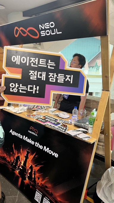 A promotional booth for NEO SOUL ('Agents Make the Move') with a man holding a speech bubble sign in Korean reading '에이전트는 절대 잡들지 않는다!' (Agents never sleep!), with merchandise and brochures on display.