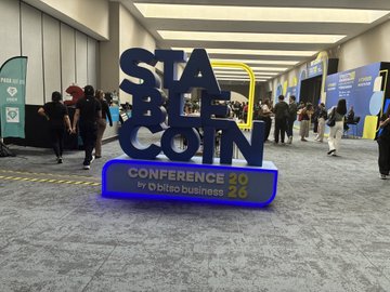 The image features an advertisement for an event held at Bitso Business, with a large blue sign displaying "Stable Coin Conference 2016" written in white letters. The conference is being advertised as a product of @bitso business, showcasing their expertise in Latin America and beyond. In addition to the prominent logo, several people can be seen walking around in front of a stage that has a "Conference" banner displayed on it, indicating the ongoing nature of the event. Overall, the image captures an exciting atmosphere at the conference and the company's involvement in promoting their product, Stable Coin.