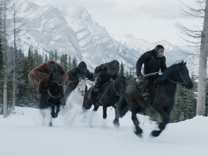 A scene from a movie showing apes and a human riding horses through a snowy landscape with mountains in the background.