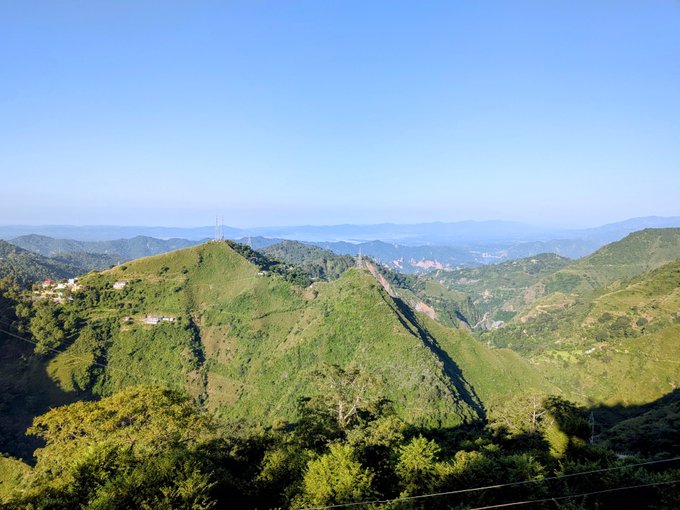 A scenic landscape view of lush green rolling mountains and hills under a clear blue sky, possibly showing the Great Wall of China in the distance.