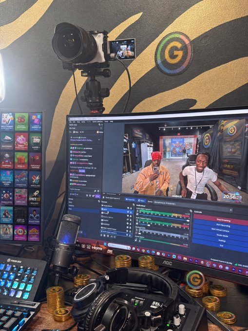 A streaming/recording setup featuring a camera on a tripod, an AOC monitor showing broadcasting software with two people on screen, a Blue Yeti microphone, headphones, and gold coins scattered on the desk, with a 'G' logo on the wall.