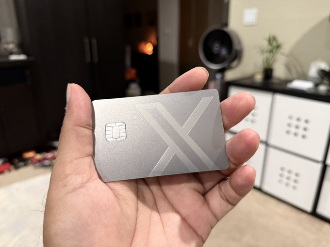 A hand proudly displays a sleek silver X-branded credit card, matching the X Money card featured in the post.