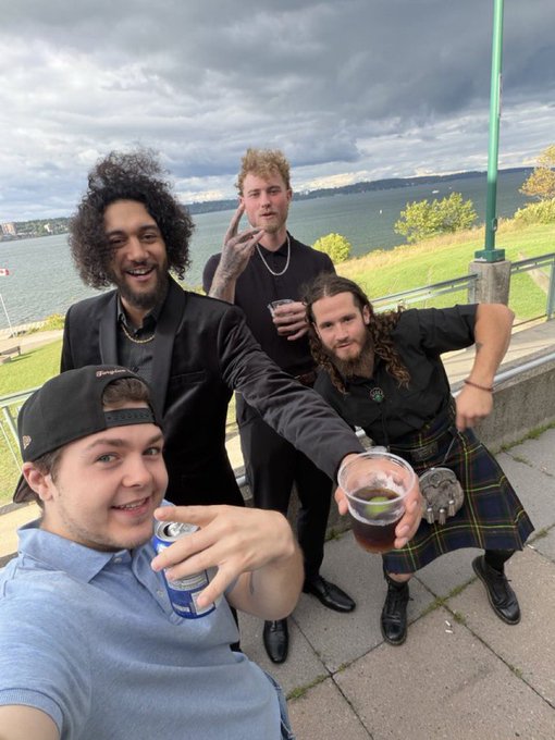 A group selfie shows four young men posing outdoors near a waterfront, with one in a kilt holding a cup, and another in the foreground holding a beer can.