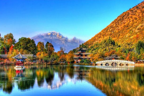 A scenic landscape featuring traditional Chinese architecture with pagodas and an arched bridge reflected in a lake, framed by mountains with snow-capped peaks and vibrant autumn foliage.