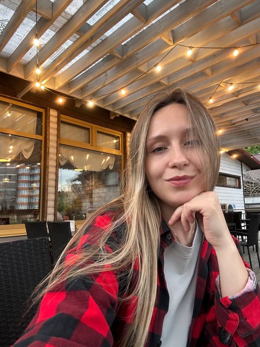 A selfie of a young woman with long blonde hair wearing a red and black plaid shirt, posing under a wooden pergola strung with lights.