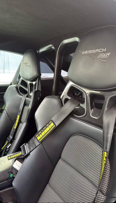 The image shows the interior of a high-end sports car featuring carbon fiber racing seats with the "WEISSACH RS" logo embroidered on the headrests and yellow Schroth racing harnesses visible. The caption "GM to everyone who says GM" humorously mocks critics with a flex of Porsche's premium, track-focused Weissach package—a top-tier option that makes the "GM" (good move) self-evident to any naysayer.