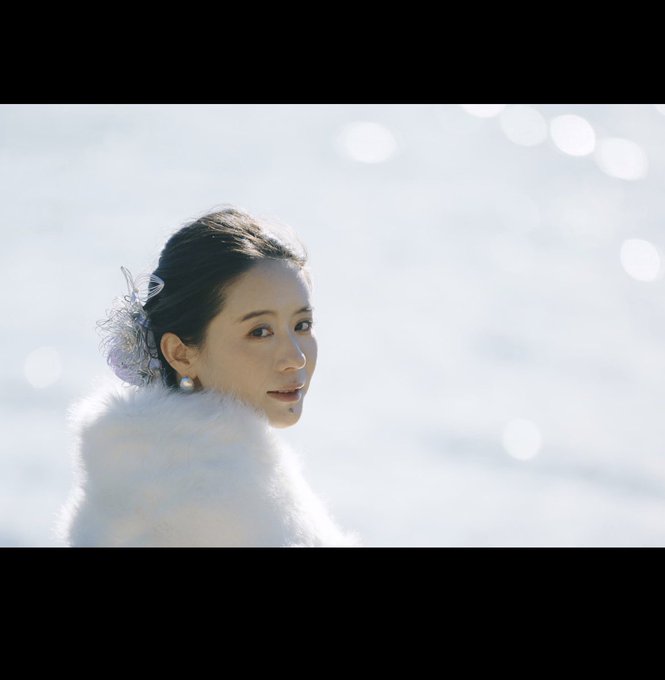 A portrait of a woman with dark hair pulled back, wearing pearl earrings and a white furry garment, set against a bright, sparkling background.
