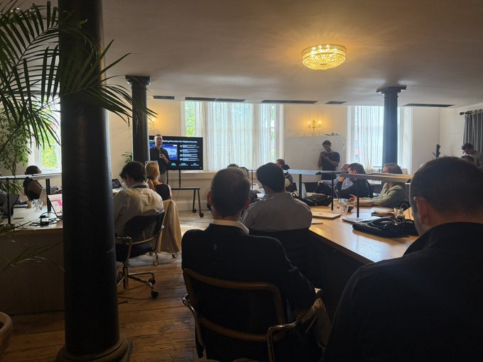 People seated at tables watching a presentation displayed on a screen in a bright room with columns, a chandelier, and a presenter standing in the background.