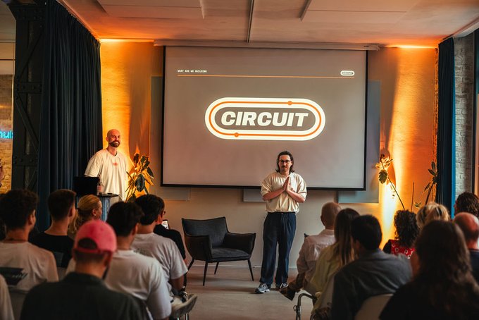A photo of a presentation or event featuring two men standing on a stage in front of an audience, with a screen displaying a 'CIRCUIT' logo behind them.