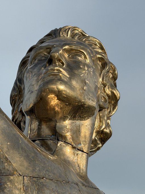 A close-up view of a weathered golden statue of a face looking upward against a pale sky.