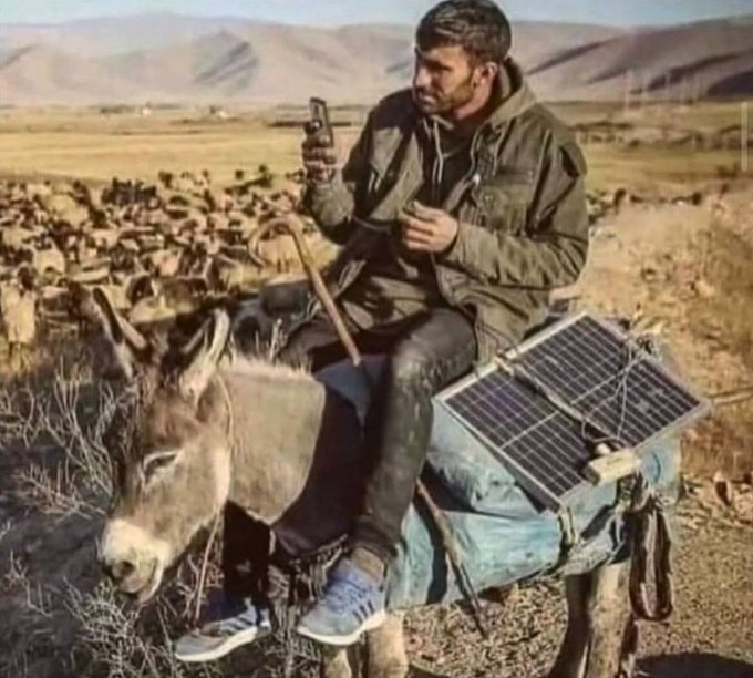 The image features a man sitting on top of a donkey, using his cell phone while riding the animal. The backdrop includes a desert landscape with mountains visible in the distance. The caption for this image reads "Crypto has no boundaries. $BTC $XPR $METAL $MTL $LOAN".