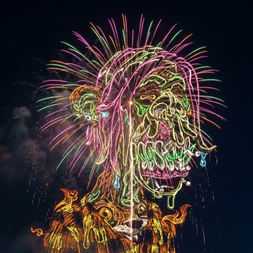 A vibrant fireworks display shaped like a screaming skull or demonic face in neon pink, green, and yellow against a dark night sky.