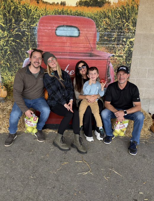 A group photo of five people sitting on hay bales in front of a red pickup truck backdrop, holding baskets of apples labeled 'Pickers' at what appears to be an apple orchard.