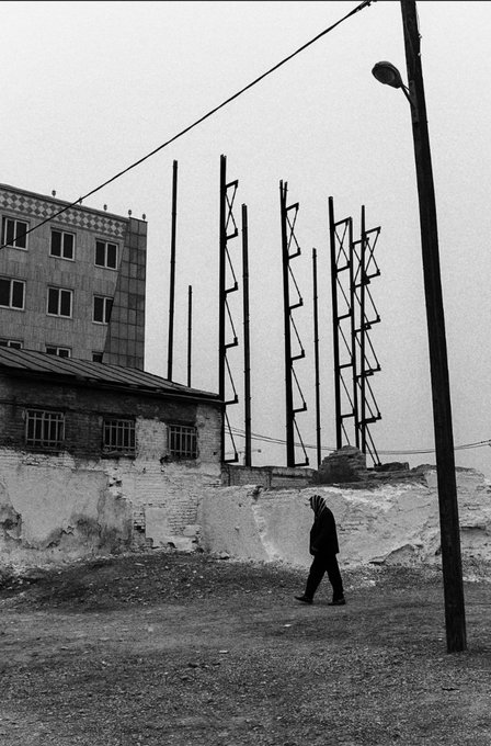 A black and white photograph shows a lone figure walking past tall metal structures and a weathered concrete wall in an industrial setting.
