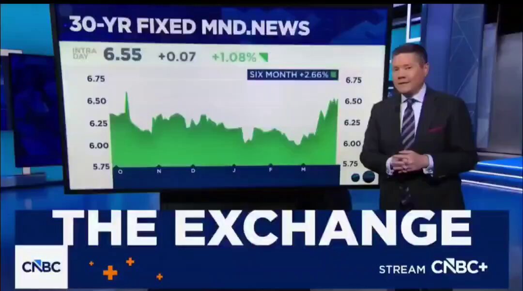 This image shows a man standing at a television set talking about the benefits of using Bitcoin as a form of payment for houses. He is wearing a suit, tie, and has a microphone attached to his mouth. The news anchor provides information on this new way of exchanging money through cryptocurrency in relation to buying homes.