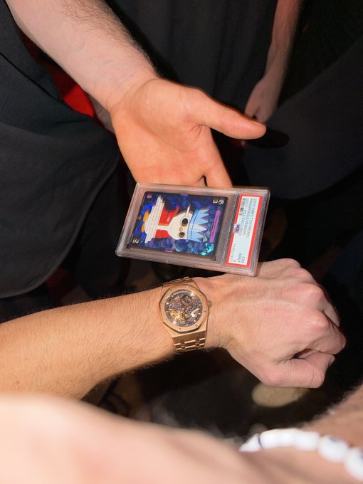 A man presents a PSA-graded Pokémon trading card (appearing to be a Mewtwo illustration rare, rated 10) alongside another person's rose gold skeleton watch, capturing a moment from a NYC happy hour where fellow collectors showcase their prized possessions.