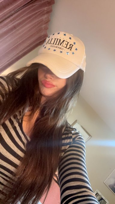 A woman with long dark hair wears a cream-colored baseball cap with 'ESSENTIALS' written backwards and a black and white striped long-sleeve shirt.