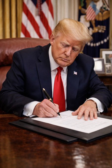 President Donald Trump is shown at his desk in the Oval Office, focused on signing a document, set against the backdrop of the American flag—symbolizing official presidential action on policy matters, including his recent remarks endorsing the growing use of Bitcoin as a positive development for the country.