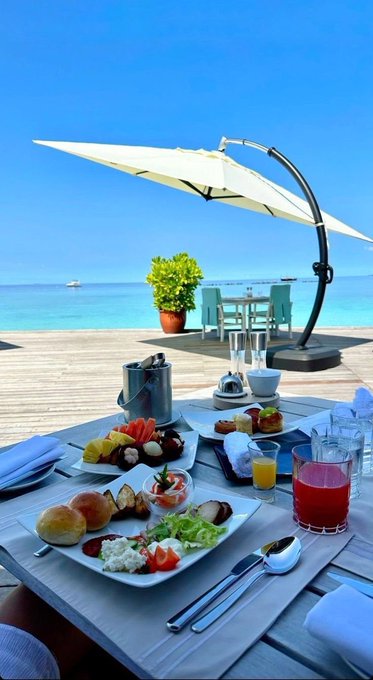 A luxurious beachside breakfast spread with fruit, pastries, salads, and drinks sits on a table under an umbrella overlooking turquoise water.