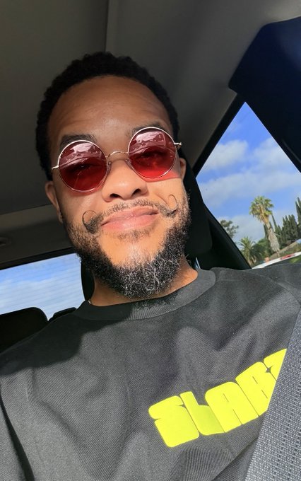 A man wearing round red-tinted sunglasses takes a selfie inside a car, sporting a black t-shirt with the yellow "SLABZ" logo. The image ties into the post's promotion of the Slabzapp affiliate program, encouraging sign-ups with the discount code "REDIC."