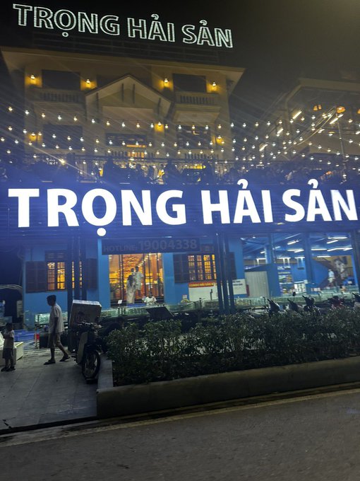 A nighttime photo of a seafood restaurant with blue illuminated signage reading "TRỌNG HẢI SẢN" and "HOTLINE: 19004338", decorated with string lights, showing a few patrons near the entrance.