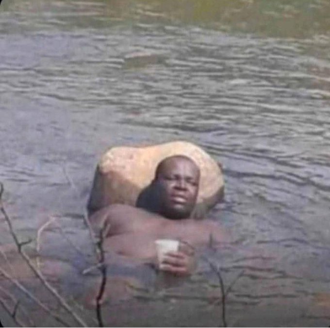 The image shows a man sitting in a body of water with his head above the surface. He has a cup in his hand, which could be filled with beer or another beverage. The background features a river flowing through a forested area, creating a serene and natural setting for the scene.