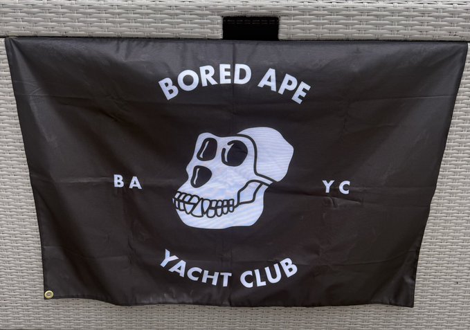 A black flag featuring the white Bored Ape Yacht Club skull logo with the letters "BAYC" and the text "BORED APE YACHT CLUB" displayed against a woven background.