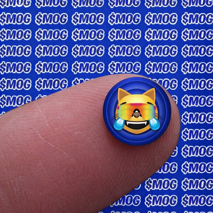 A close-up photo of a small, round pin featuring a crying, rainbow-faced cat character, held against a blue background covered in '$MOG' text.
