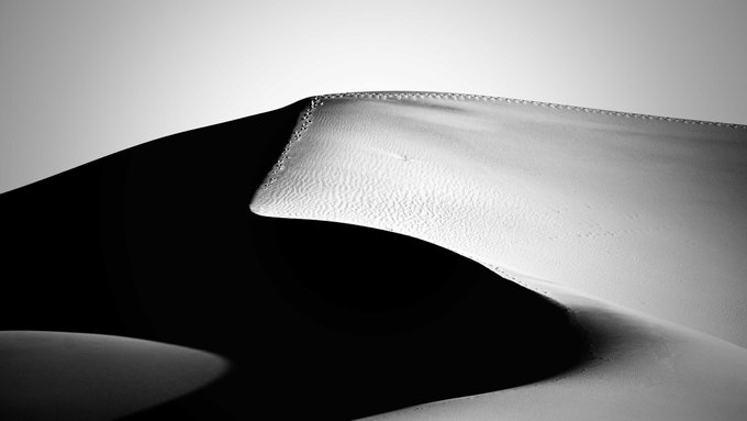 A black and white abstract image features a smooth, flowing form resembling folded paper or liquid metal with sharp edges and shadows.