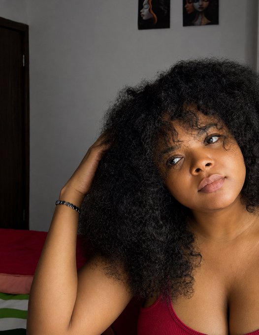 A young woman with voluminous curly hair poses in a red top, looking off to the side in a softly lit room with artwork on the wall behind her.