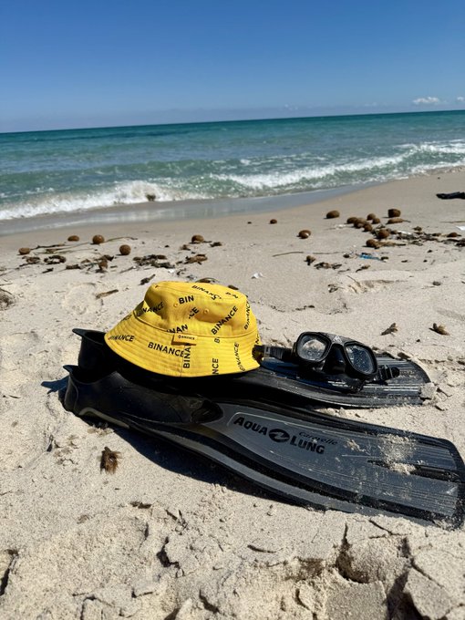 Snorkeling gear—a yellow "Binance" bucket hat, "Aqua Lung" fins, and a dive mask—rests on a sandy beach, perfectly capturing a laid-back Friday by the shore.