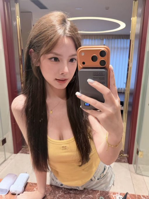 A young woman takes a mirror selfie with her iPhone, with "HAMI00001" visible on a card attached to her phone case, as she preps for her trip to TOKEN2049 Singapore and KBW. The selfie captures a casual, travel-ready moment before her Asia tour focused on meeting KOLs, traders, and founders in the crypto space.