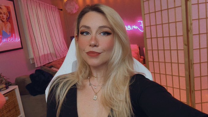A blonde woman with red lipstick wearing a black top and necklace poses for a selfie in a pink-lit room, with a Marilyn Monroe poster on the left wall and a neon sign in the background.