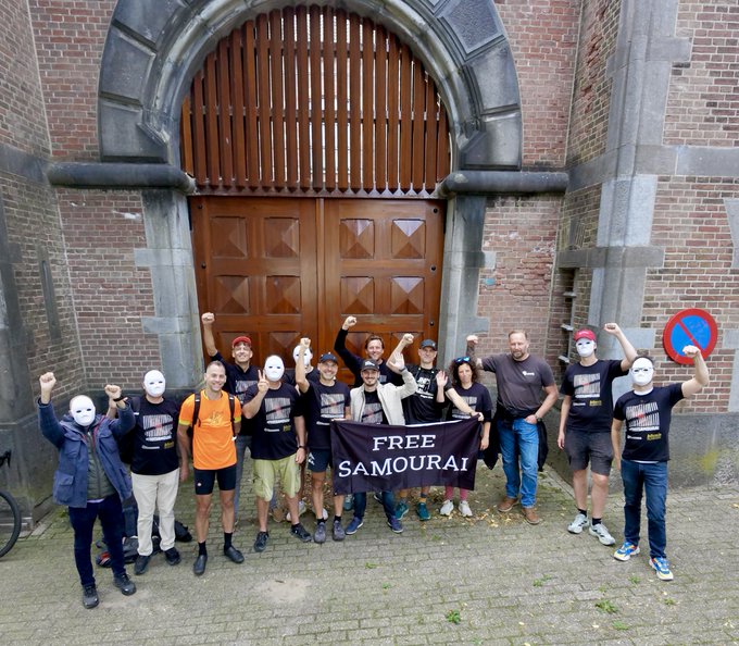 A group of people wearing black t-shirts and white Pepe frog masks holding a 'FREE SAMOURAI' banner in front of a brick building.