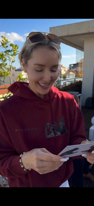 The image features a woman wearing a red hoodie, smiling as she reads an email from someone named @XCOPYART. The woman appears to be engaged with the content of the message, possibly discussing or sharing information related to the email. The background shows a building and trees, suggesting that they might be in an outdoor setting like a park or garden.