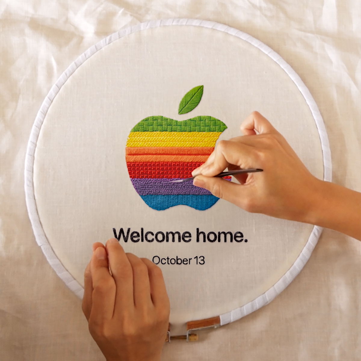 Two hands work on an embroidery hoop featuring the classic rainbow-striped Apple logo with the text 'Welcome home.' and 'October 13' below it.