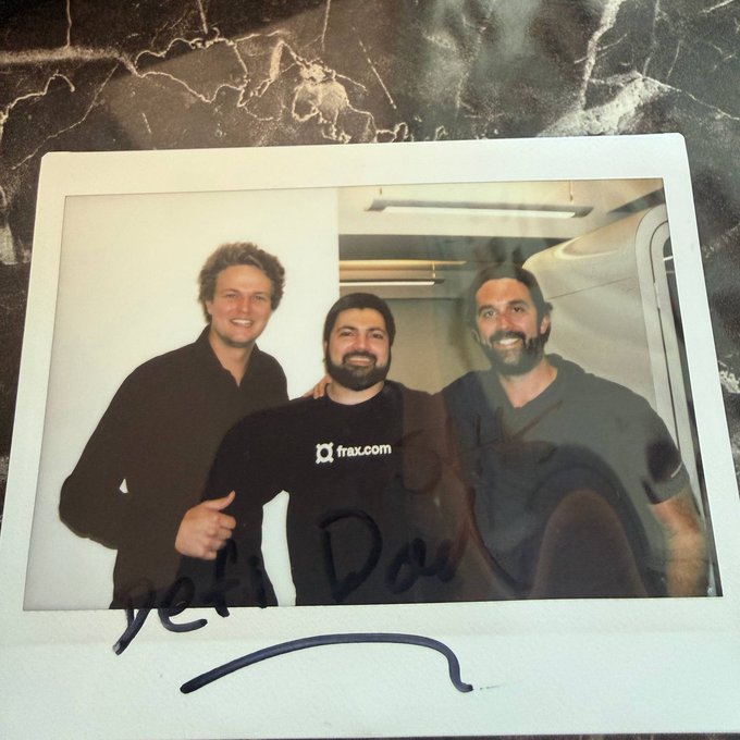 The image features three men posing for a picture, all wearing black shirts. The man in the center has his hand raised in a thumbs up gesture, while the other two men are smiling at him. The background of the photo shows a marble countertop with a glass window behind them. There is also a post-it note on the back of the image that reads "ETHConf Day 1 vibes with two GOATs @samkazemian @DeFi_Dad".